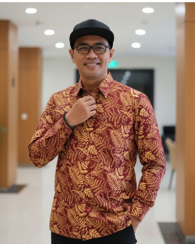 Foto Bro Trian mengenakan kemeja batik merah-emas motif pakis, topi hitam, dan kacamata